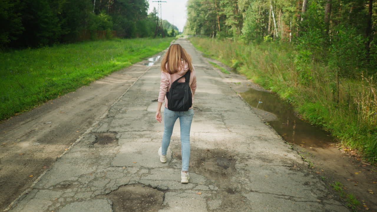 Back view of girl walking along worn tarred road with visible potholes and roadside puddles, surrounded by green grass and forest trees, wearing backpack and casual outfit under daylight sky