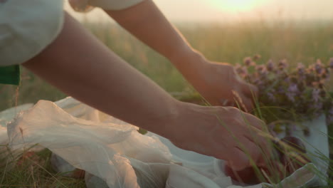 side view of woman arranging fruit and wildflowers on picnic cloth during golden hour, hands gently placing items on grass as soft sunset light creates peaceful atmosphere in warm outdoor setting