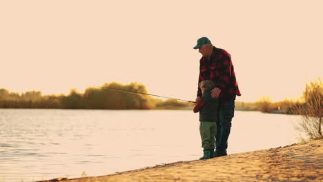 El-Viejo-Pescador-Le-Está-Enseñando-A-Su-Nieto-A-Pescar-En-Un-Lago-O-Río-Al-Atardecer,-Descanso-Activo-En-El-Fin-De-Semana.