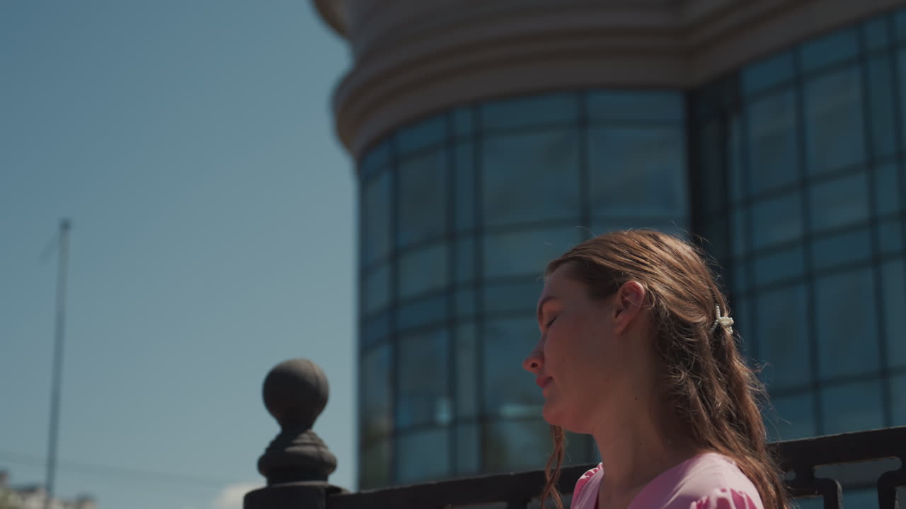 Young caucasian woman reading letters outdoors on sunlit urban terrace, pink dress with braided hair, flipping paper pages and pausing to gaze at modern glass building skyline warm film tones, gentle