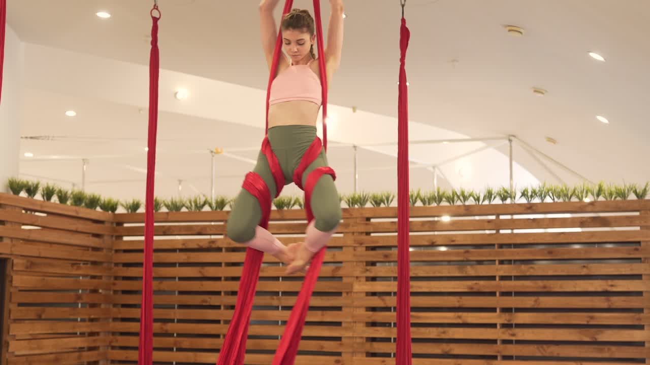 Giovane donna bruna in una palestra minimale e moderna con divisori in legno che pratica yoga aereo con alcune amache rosse brillanti, eseguendo acrobazie sorprendenti e acrobatiche.