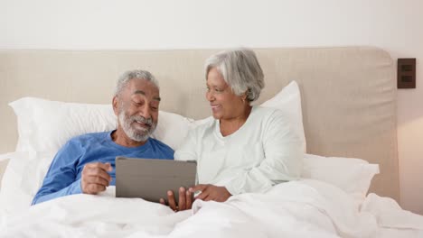 Feliz-Pareja-Birracial-De-Ancianos-Acostada-En-La-Cama-Y-Usando-Tableta,-Inalterada,-Espacio-Para-Copiar,-En-Cámara-Lenta