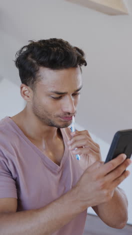 vertical video of latino man brushing teeth and using smartphone in bathroom