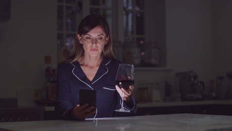 mujer infeliz vestida con pijama sentada en la cocina con un vaso de vino por la noche usando un teléfono móvil