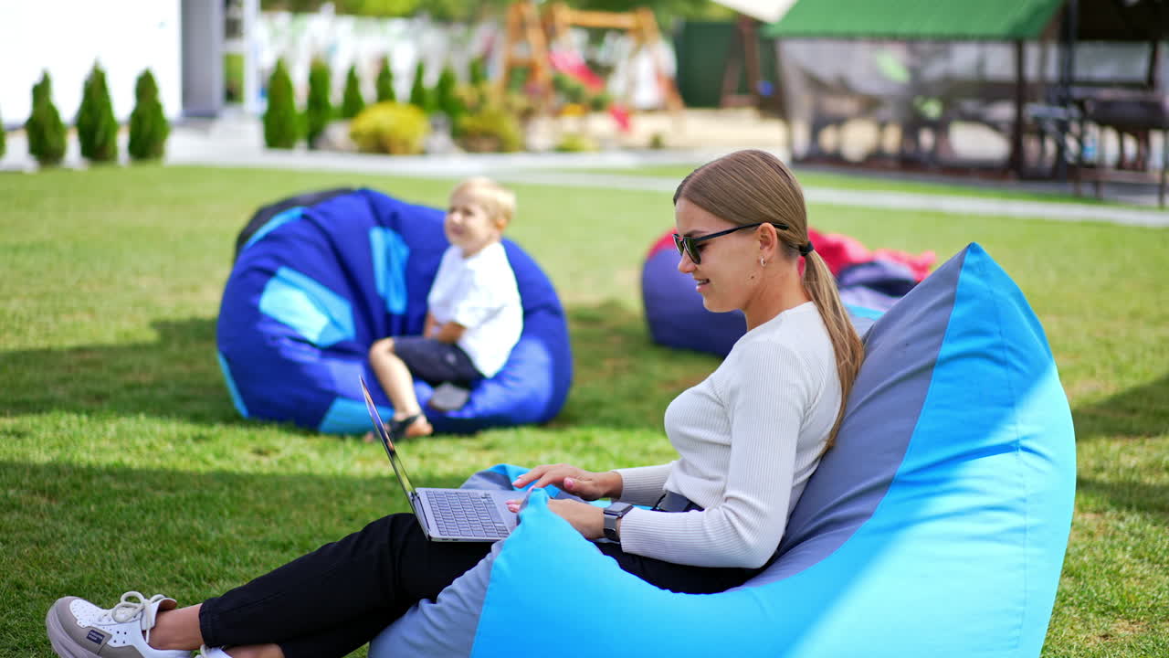 Young blond lady in sunglasses works at laptop sitting in bean bag chair. Her son sitting not far from mother saying something. Blurred backdrop.