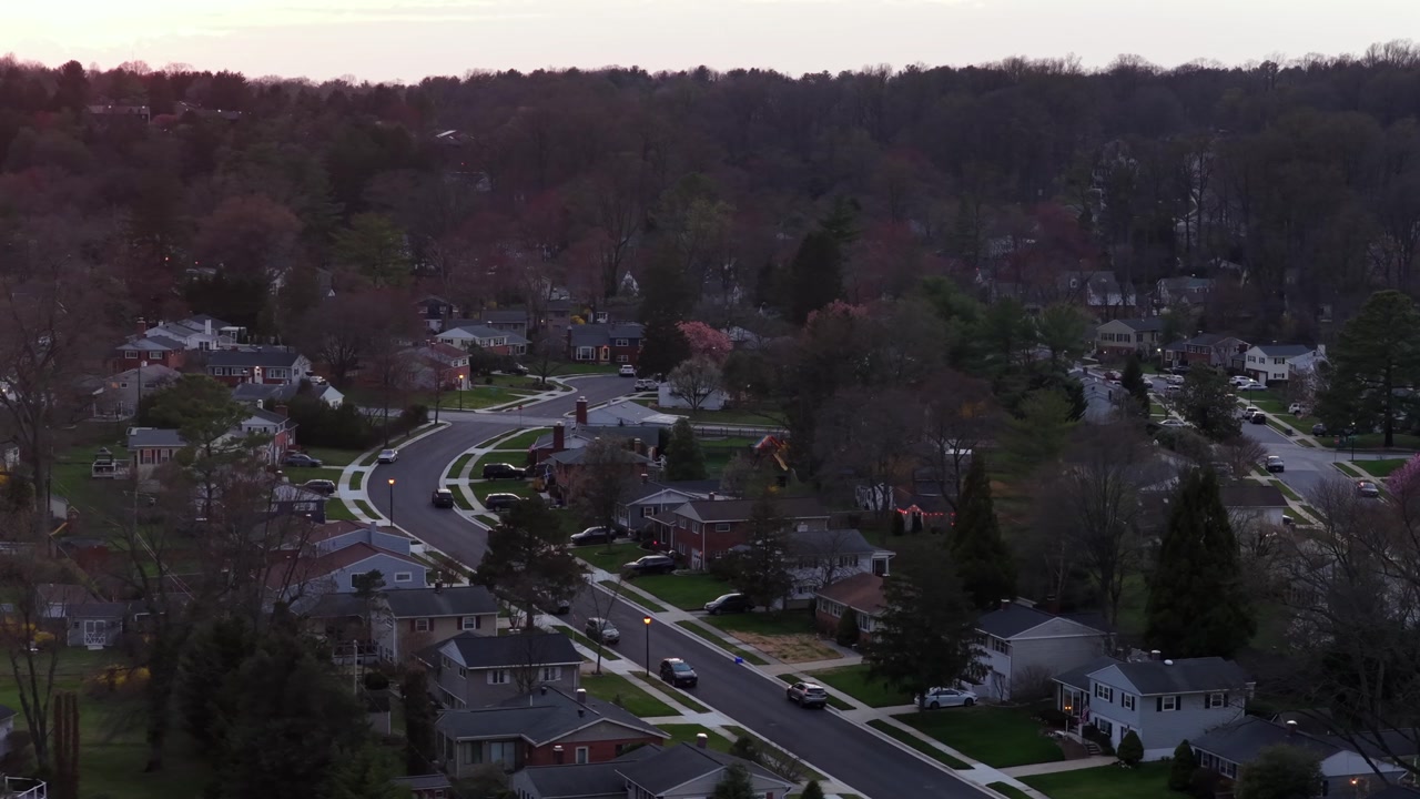 Wide aerial view of quiet suburban neighborhood with tree-lined streets and residential homes, captured in soft spring light with early foliage and calm atmosphere. Orbit shot.