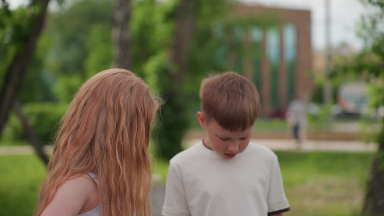 Youngsters looking down with focus on ground, blurred foliage of recreational center behind, quiet moment of shared curiosity and subtle conversation during outdoor play in summer green setting