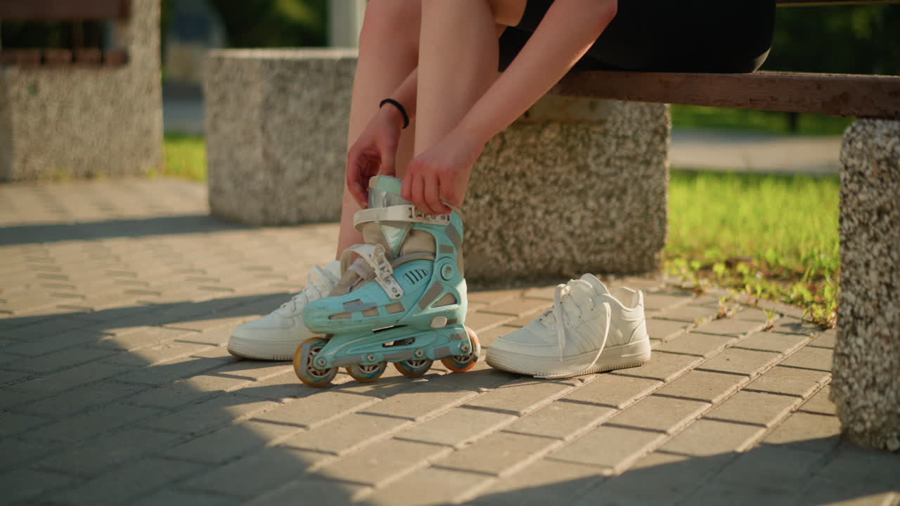 Side view of individual seated on bench wearing roller skate on left leg while right leg wears sneaker, with sneaker placed nearby, sunlight casts warm shadows on interlocked pavement