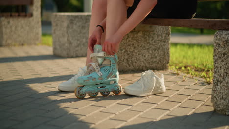 side view of individual seated on bench wearing roller skate on left leg while right leg wears sneaker, with sneaker placed nearby, sunlight casts warm shadows on interlocked pavement