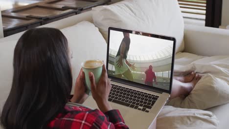 Composite-of-woman-sitting-at-home-holding-coffee-watching-rugby-match-on-laptop