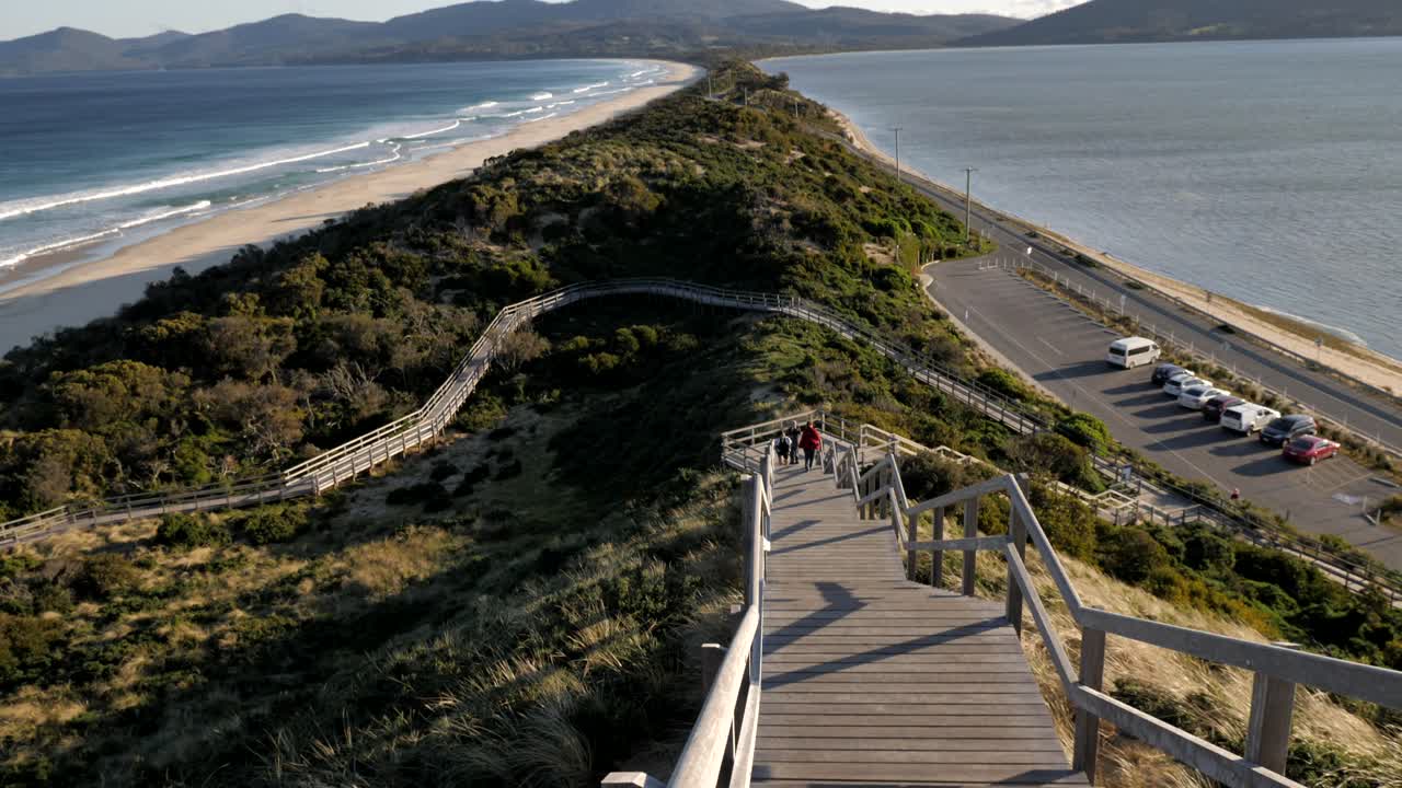 산들바람이 부는 봄 저녁 동안 배경 해안선 숲 산에서 굴러가는 파도와 함께 호주 브루니 섬 태즈메이니아에 있는 목 지협 나무 목재 보드워크 위로 천천히 팬