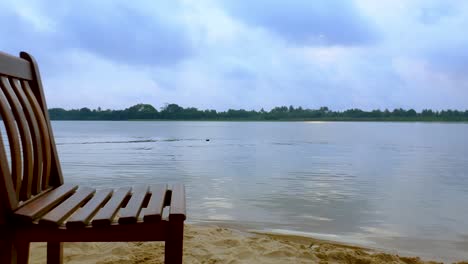 Silla-De-Madera-Vacante-A-Lo-Largo-Del-Río-Silencioso-Y-Tranquilo-En-Un-Día-Nublado