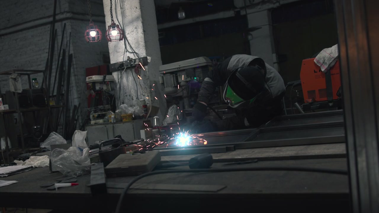 Single worker in protective welding helmet bends over metal fabrication bench inside dim industrial workshop, with bright welding arc, sparks, cluttered tools, pipes, cables, drill press, concrete