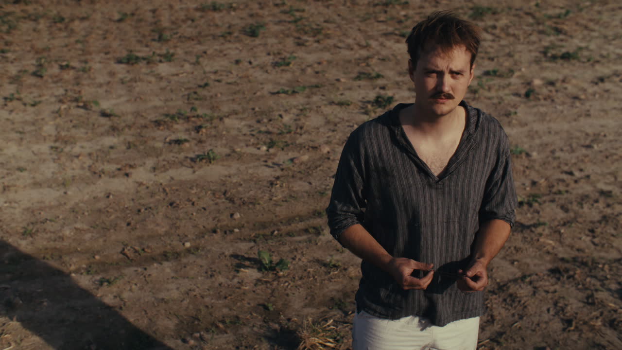 A man stands in a sparse field, looking down at his hands