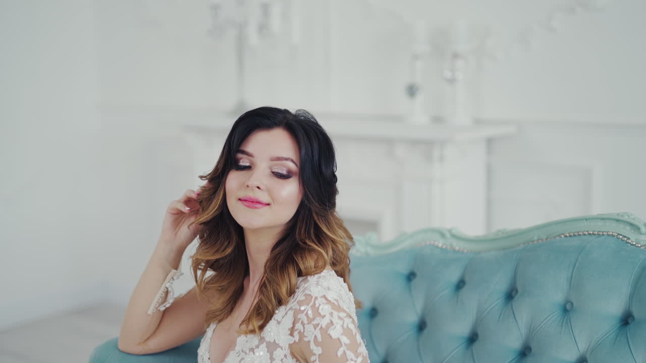 Incredible young woman in a beautiful white wedding dress is sitting on the couch. Lovely model with delicate makeup posing on royal sofa and smiling to camera.