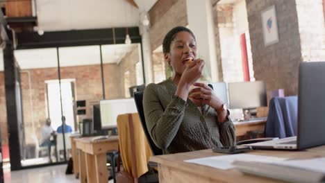 happy african american casual businesswoman using laptop and having lunch in office, slow motion