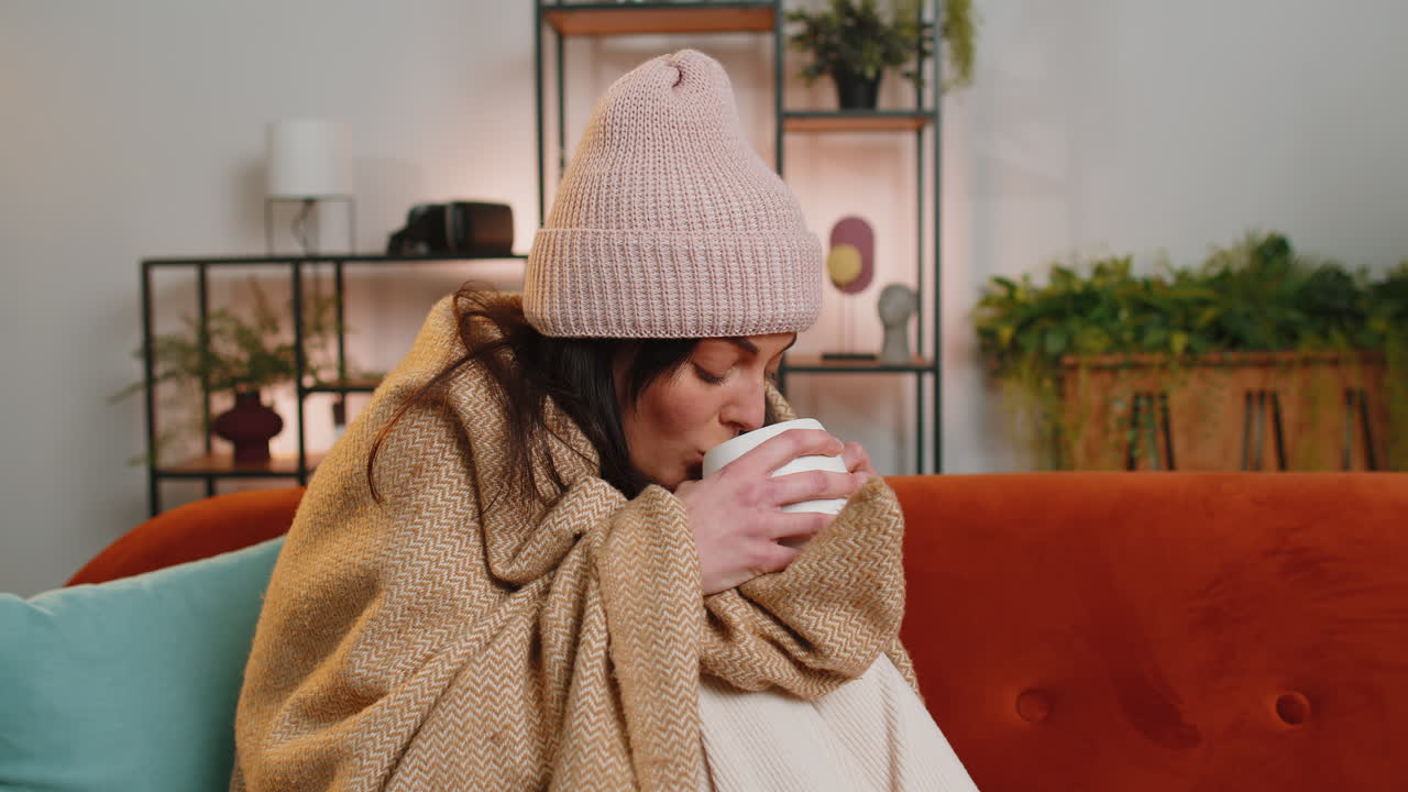 Sick unhealthy woman wear hat wrapped in plaid sitting shivering from cold on sofa drinking hot tea