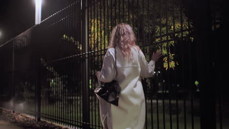 lady in white coat carrying black handbag and umbrella walking along sidewalk at night touching iron fence illuminated by soft streetlights with scattered autumn leaves on ground