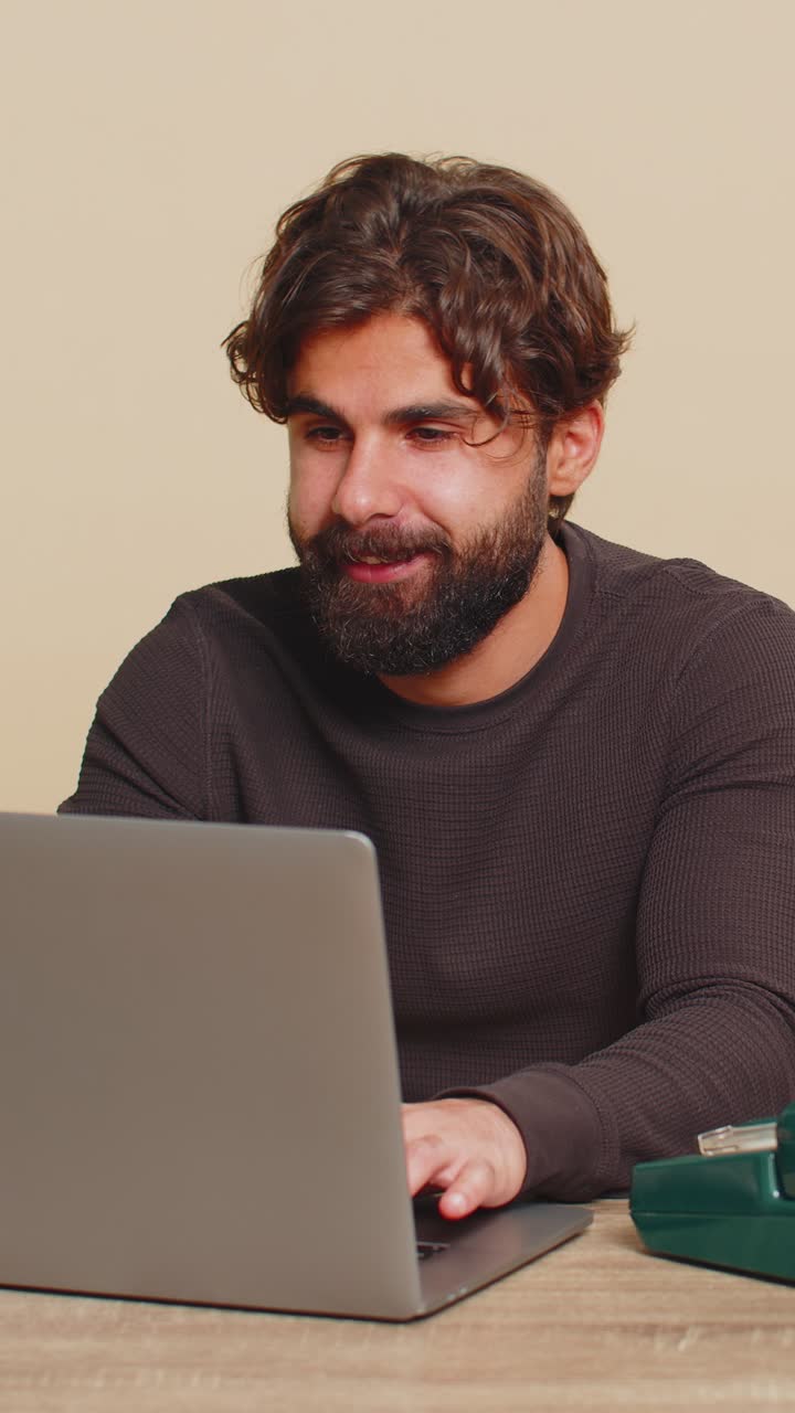 Happy businessman sitting at table talking on wired telephone and using laptop on beige background