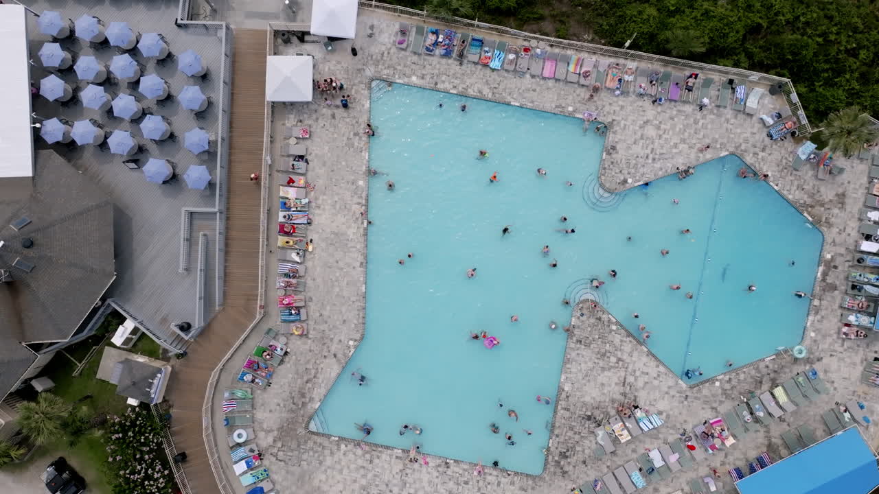 Ovenfra-udsigt af en stor geometrisk pool fyldt med badegæster, omgivet af liggestole, parasoller og nærliggende resortbygninger