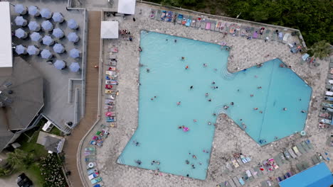 top-down view of a large geometric pool filled with swimmers, surrounded by loungers, umbrellas, and nearby resort buildings