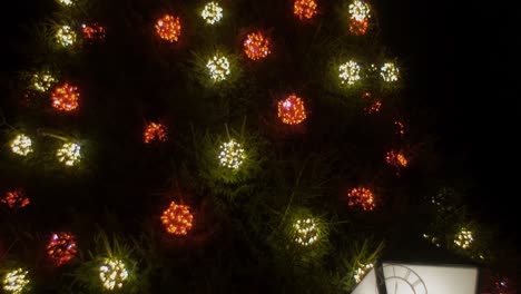 Weihnachtsbaum-Im-Freien,-Geschmückt-Mit-Blinkenden-Lichtern-Und-Weihnachtsmütze-Auf-Der-Spitze-Des-Baumes,-Laterne-Mit-Uhr-Overlay,-Nach-Oben-Schwenkbare-Kamera