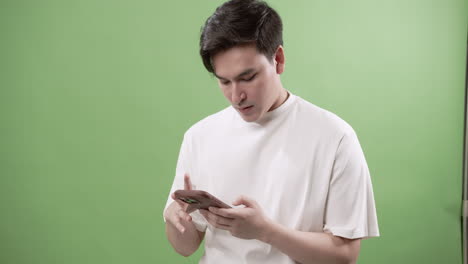 A man is standing in front of a green background holding his mobile while browsing the internet.