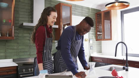 Happy-diverse-couple-standing-in-kitchen,-using-tablet-and-rolling-dough,slow-motion