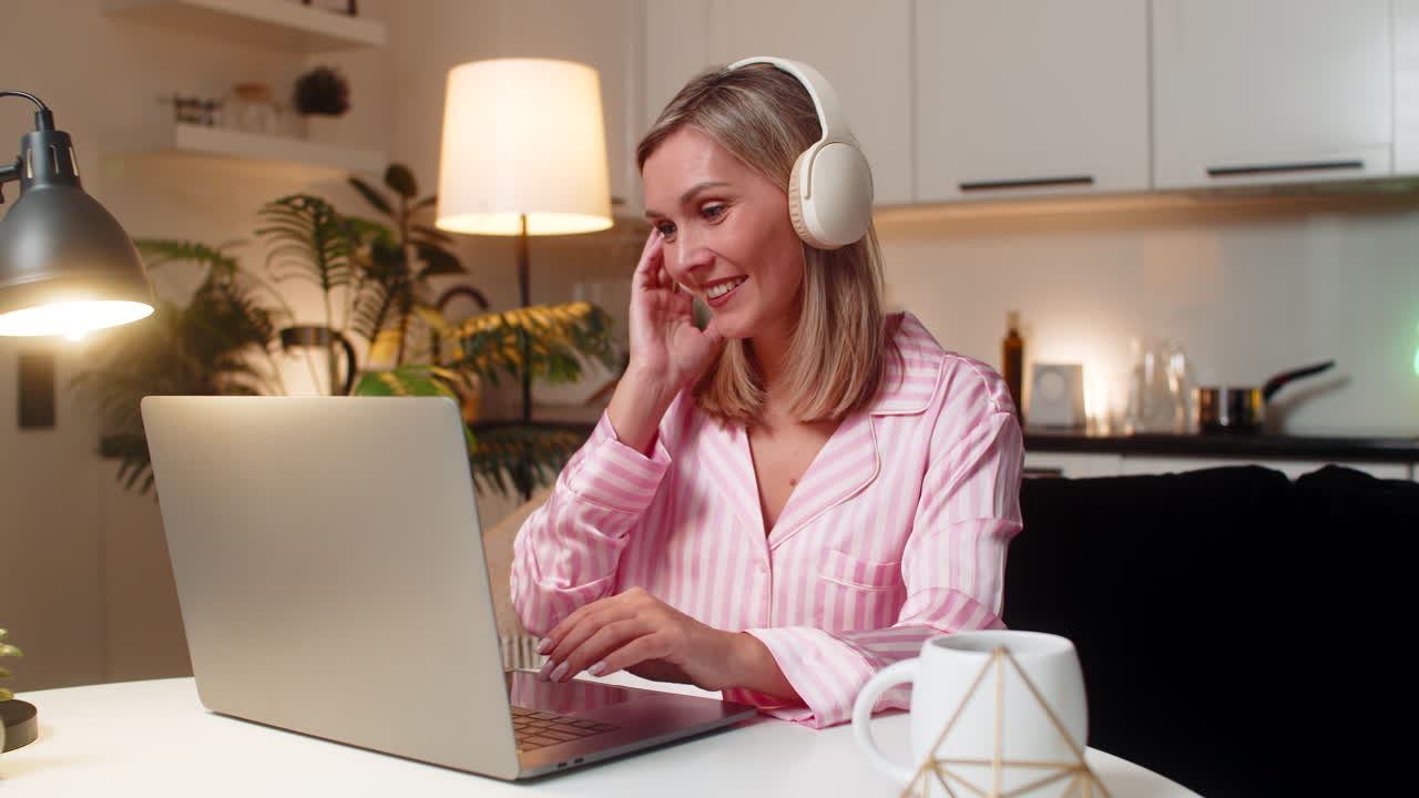 Voksen kvinde med hovedtelefoner justerer layout på bærbar computer, flytter lag, nikker til musikken, smiler og gemmer versionen