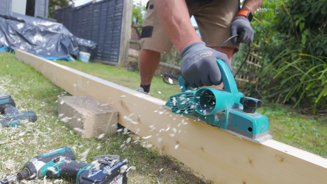 A wide shot of plank of wood being planed in a garden by a man on a sunny day in 4 x slow motion