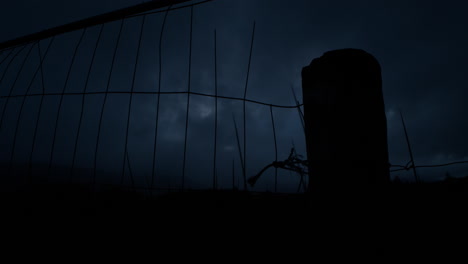 dark night sky with clouds moving, metal fence and stone fest post in the foreground, dark silhouetted grasses swaying in the wind, very moody atmosphere, a bat flying across