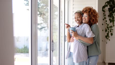 Happy-diverse-female-lesbian-couple-embracing-and-looking-through-window-at-home-in-slow-motion