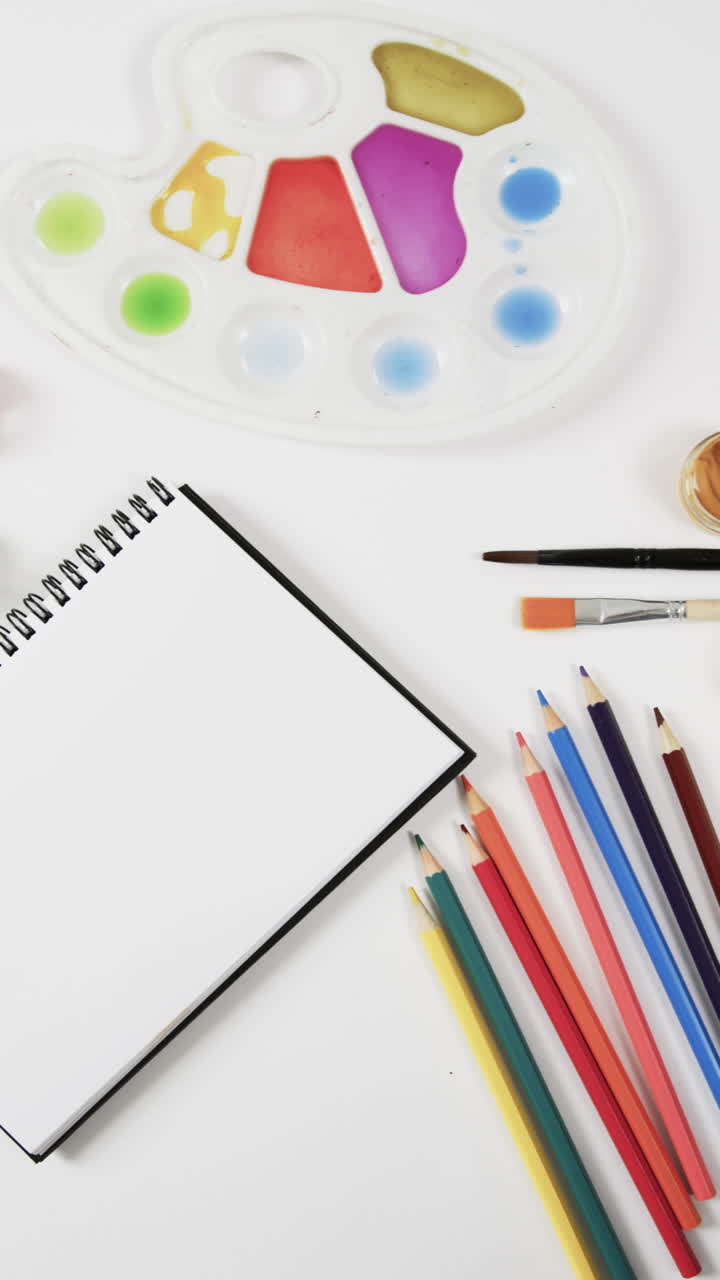 Vertical video of overhead view of notebook, paints, brushes and crayons on white background