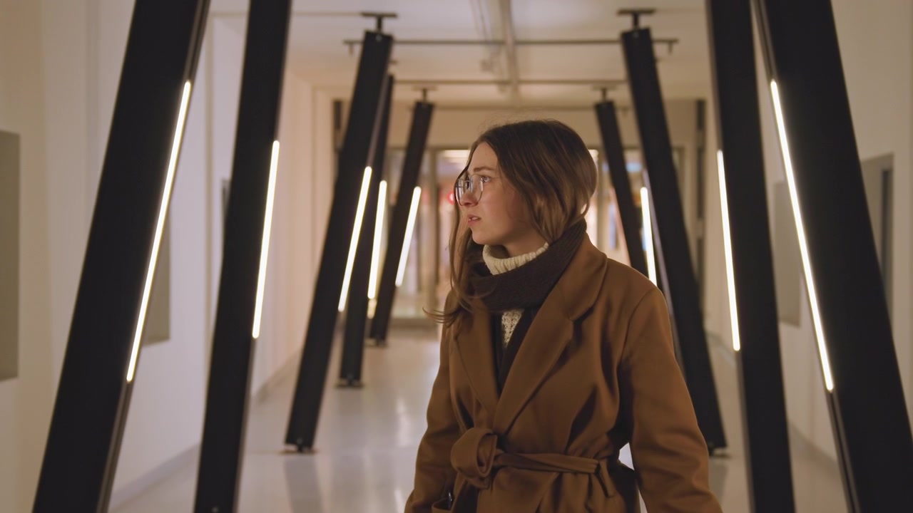 Mujer en un pasillo moderno. Caminando con un ánimo reflexivo en el interior. La mujer se mueve silenciosamente por un pasillo bien iluminado. La mujer vestida de marrón avanza por un espacio moderno con iluminación simétrica.