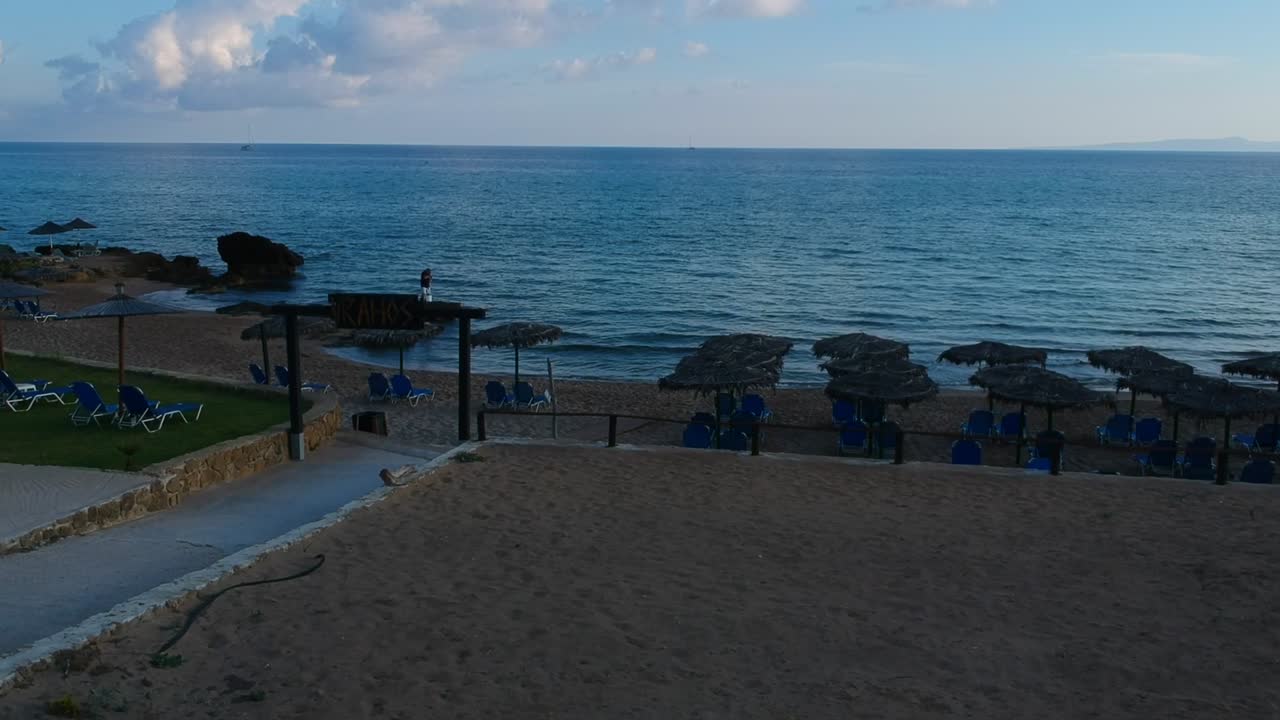 Plaka beach resort huts in the Vrahos bar sign with Man fishing on the rocky shore, Aerial drone dolly-in shot