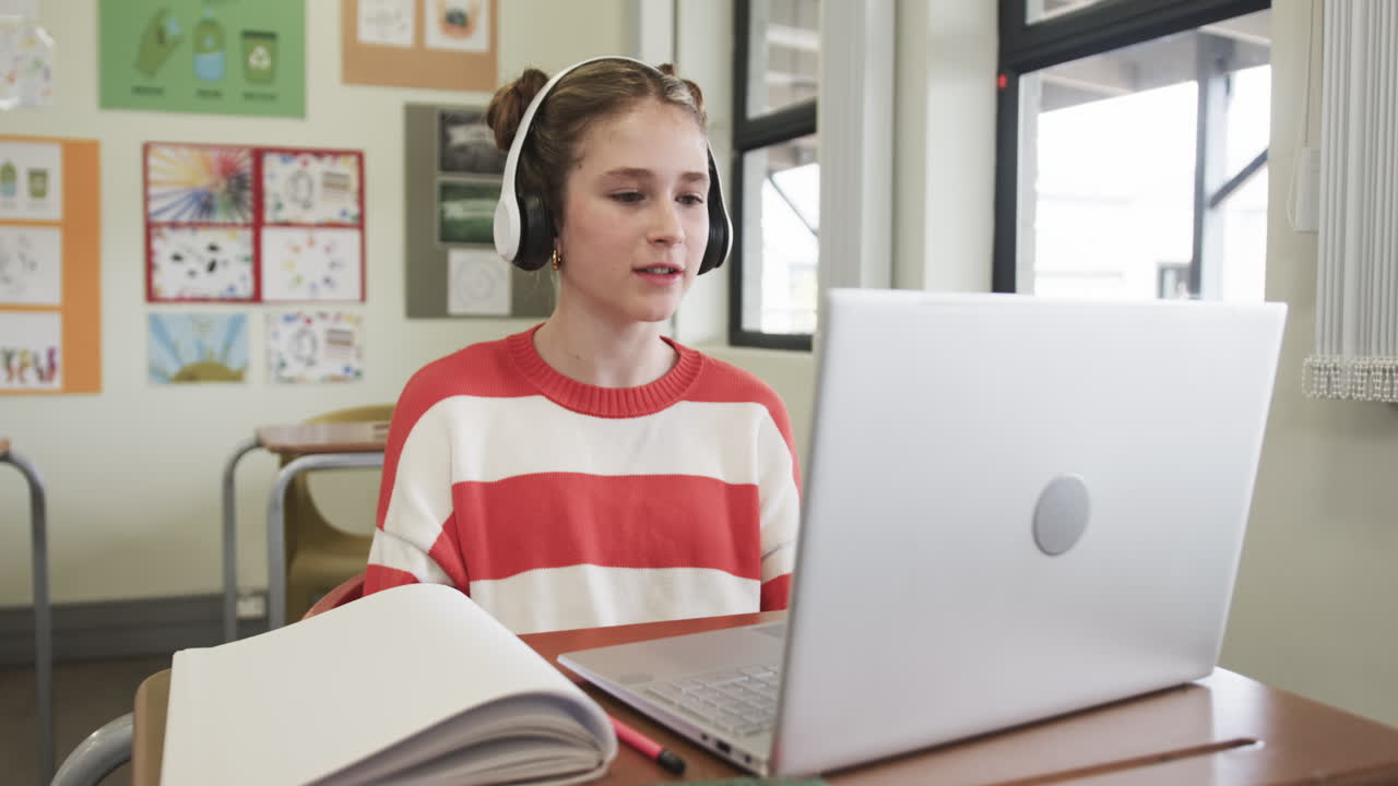 On video call, young girl wearing headphones in online class with laptop in classroom, at school