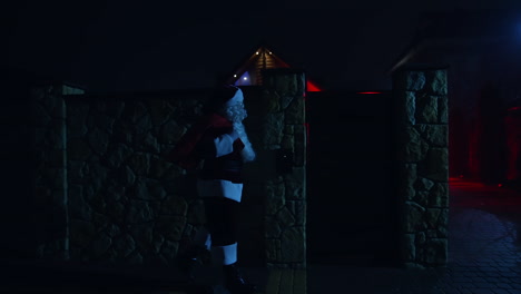 santa claus with red bag at night. santa walks into front yard of house decorated with lights and garlands. santa carrying gifts for children.