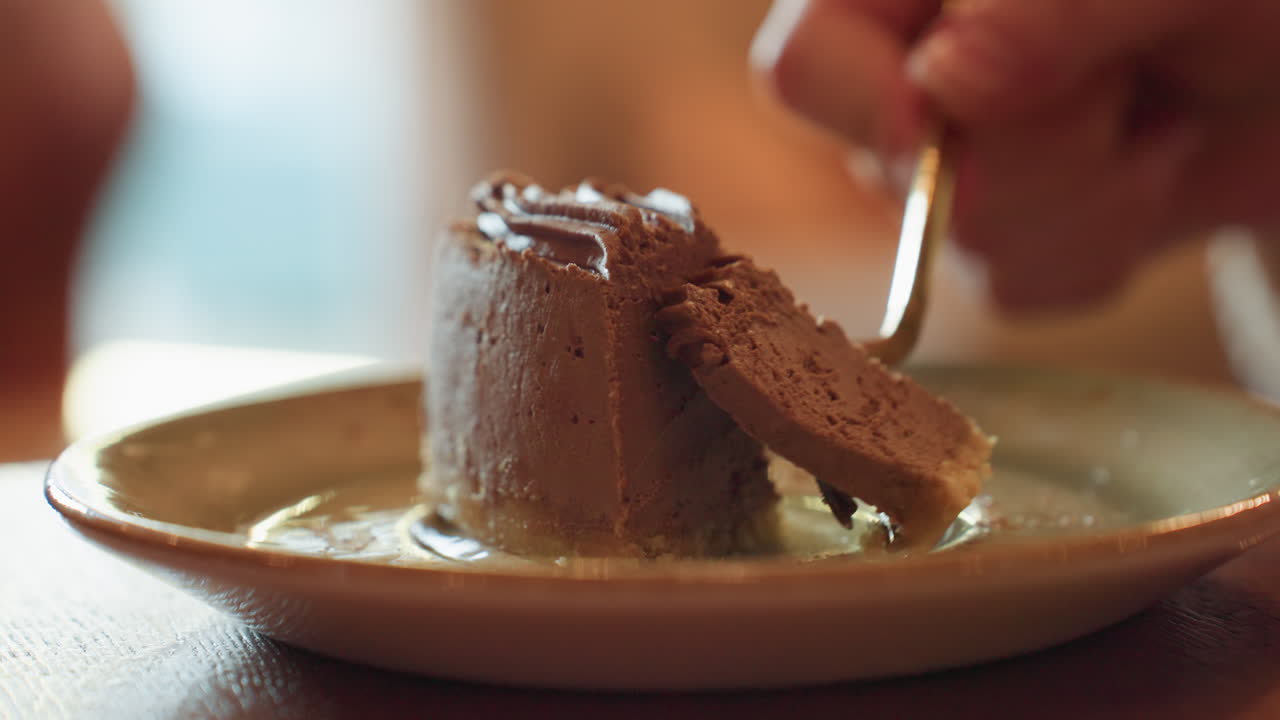 close up of hand gently slicing rich chocolate dessert with fork on ceramic plate, soft blur background adds cozy mood and highlights creamy texture and delicate detail of sweet treat in warm setting