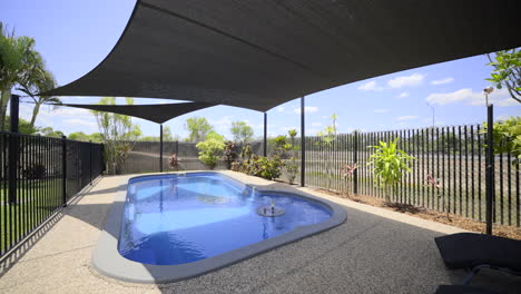 large luxurious contemporary outdoor swimming pool in ground, clear blue water black fence grey concrete decking modern home green grass blue skies dark shade sails pool chairs