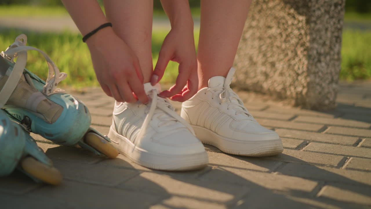 오른쪽 다리 스니커의 신발을 풀고 보행자 에 앉아있는 여자의 가까운 모습, 그녀의 에 누워있는 롤러 스케이트, 초록색과 부드러운 빛의 배경으로 땅에 던진 그림자