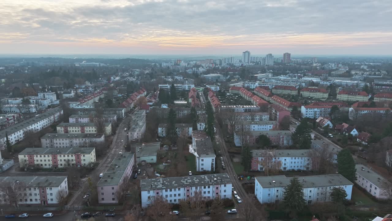Płynne ujęcie z drona przesuwające się do tyłu ukazuje rozległe przedmieścia Berlina z jednakowymi blokami mieszkalnymi i domami z czerwonymi dachami o świcie, a w oddali wyłaniają się wieżowce pod miękkim pastelowym niebem.