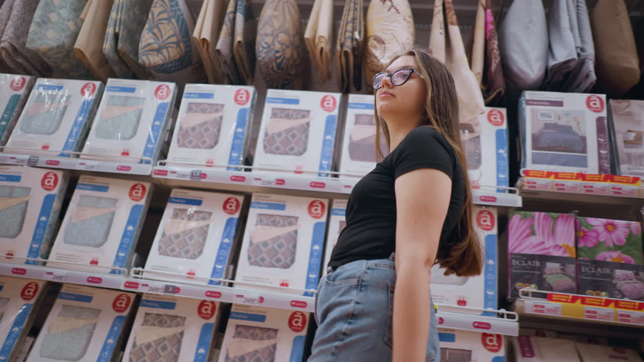 vista superior de una mujer joven comprando en una tienda, caminando por un estante con productos de cama, mira a su alrededor, observando diferentes opciones, pasillo de la tienda lleno de textiles para el hogar, almohadas y juegos de cama