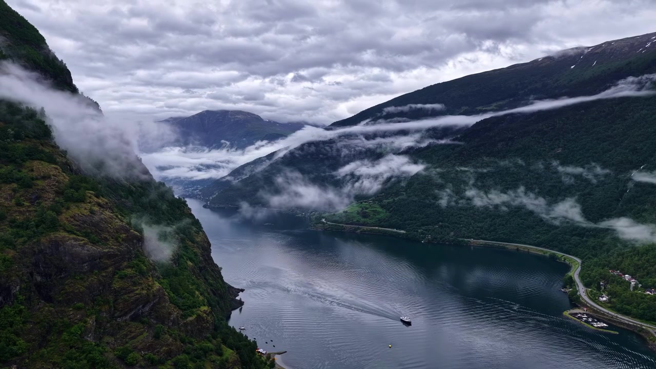 Tiefe Wolken ziehen über steile grüne Berge über einem ruhigen Fjord mit Boot in Flåm, Aurland, Norwegen.