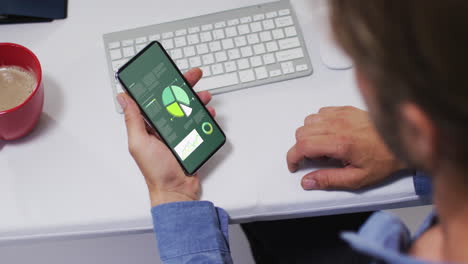 hombre caucásico sentado en un escritorio usando un teléfono inteligente con estadísticas en la pantalla