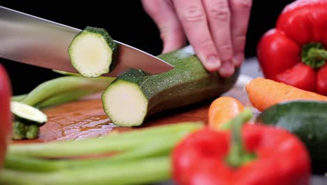 person with sharp knife cuts zuccini into slices between various fresh vegetables peppers, carrots, leeks on cutting board