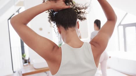 Pareja-De-Lesbianas-Birraciales-Cepillándose-Los-Dientes-Y-Peinando-El-Cabello-En-Un-Baño-Soleado,-Cámara-Lenta