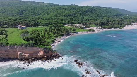 Aerial-backwards-shot-of-Playa-Quemaito-with-rocks-in-sea,-tropical-landscape-and-overgrown-mountains-in-Background---beautiful-sunny-day-on-coastline-of-Barahona