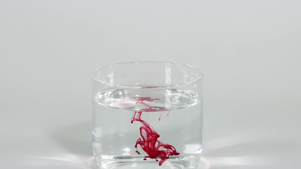 Red dye creates intricate patterns as it disperses in a clear glass of water.