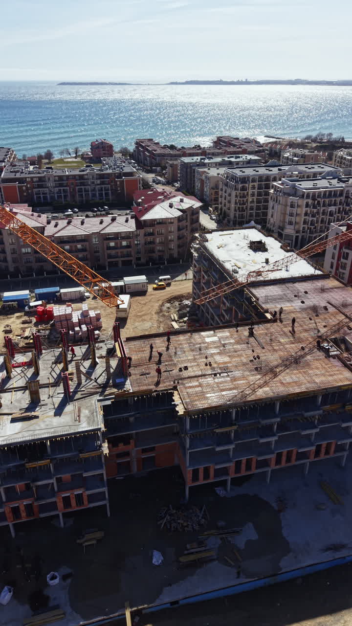 Construction workers build a high-rise building near the waterfront in bright daylight with cranes and equipment present on the site