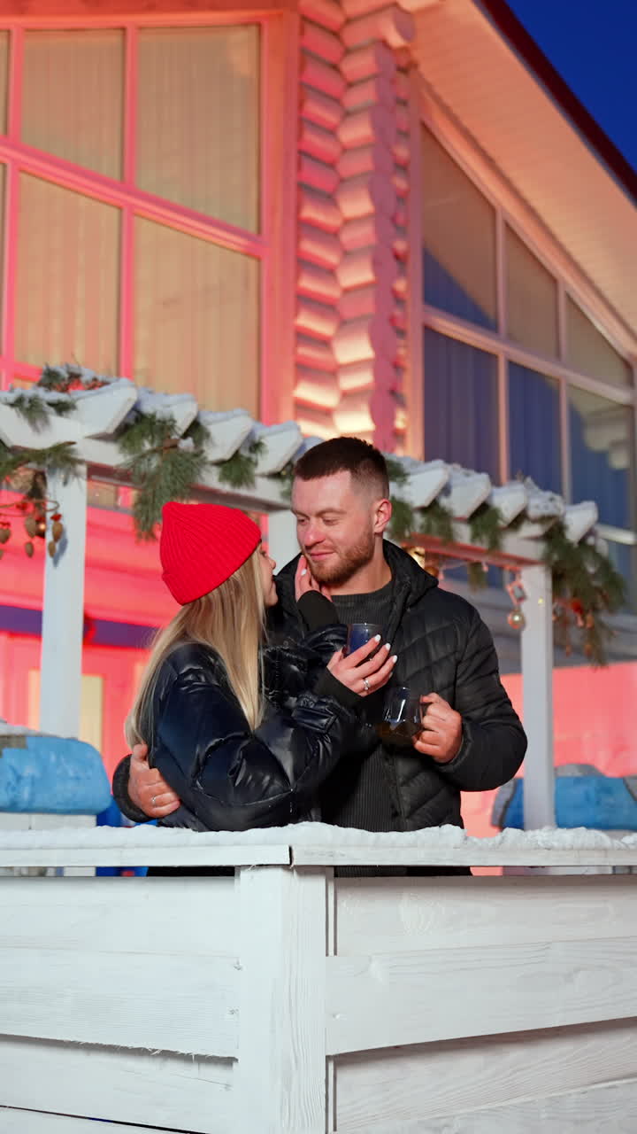 Happy woman wearing warm clothes laughs and turns to her man. Beautiful couple in love spends cozy calm evening drinking hot tea at the porch in winter. Vertical video.